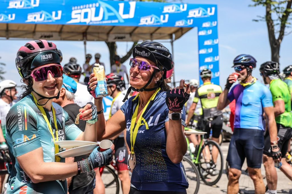 Due cicliste sorridenti con medaglia e ristoro post-gara sotto lo striscione blu della Granfondo Squali.
