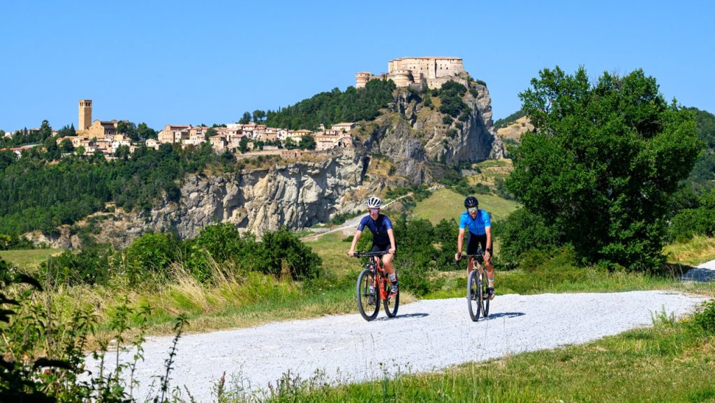 Two cyclists on gravel bikes pedaling along a white road in Valmarecchia with the majestic fortress of San Leo perched on the cliff in the background