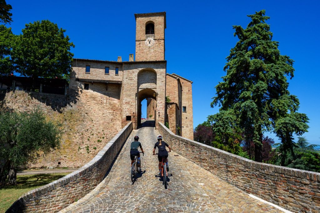 Two cyclists on gravel and mountain bikes tackling the uphill stone ramp toward the medieval gateway of the fortified village of Montegridolfo