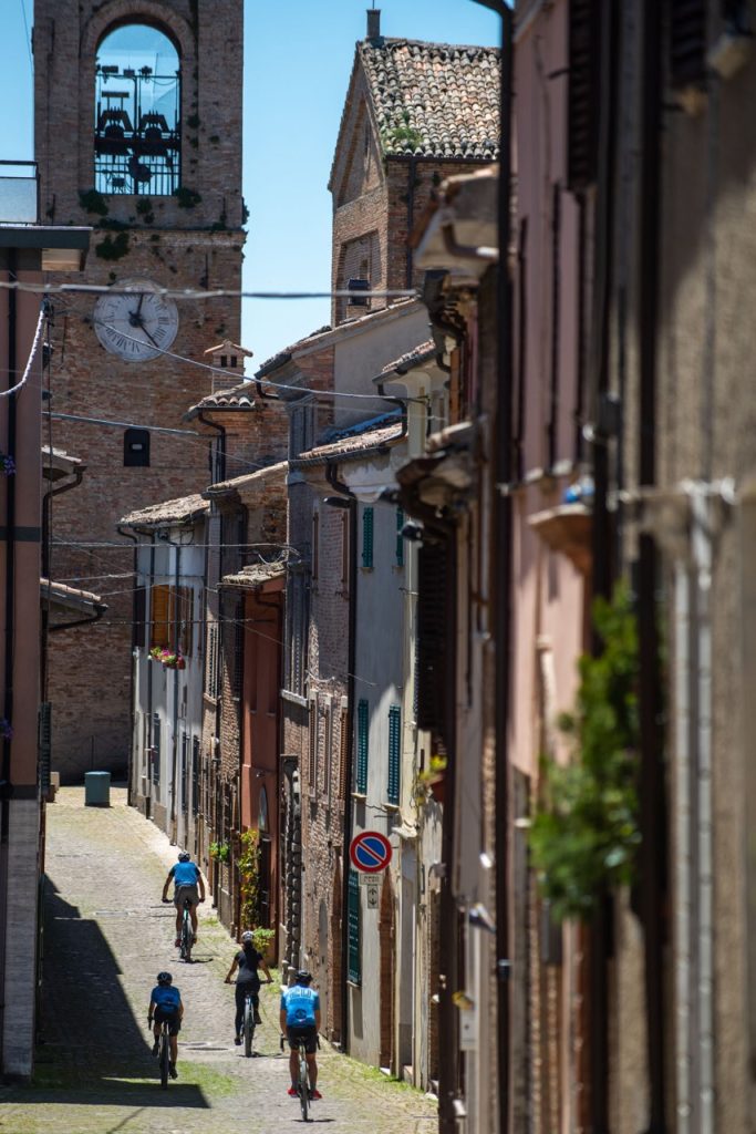 A group of cyclists riding their gravel bikes through a narrow cobblestone street in the historic medieval center of the village of Mondaino